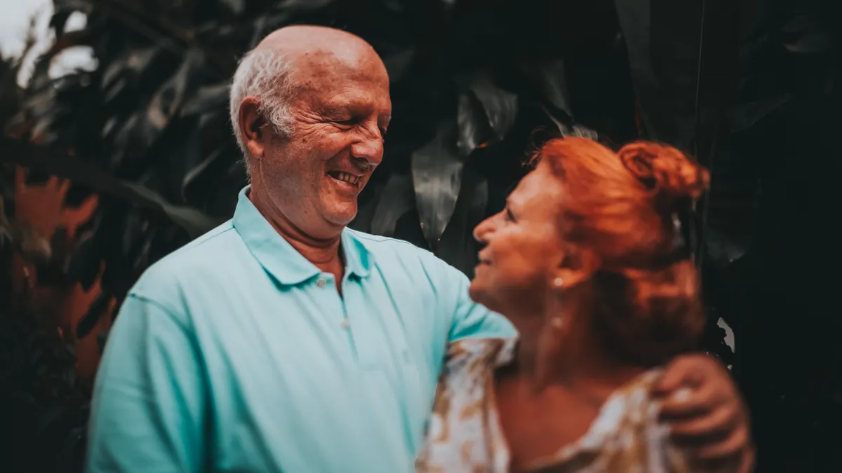 Elderly couple smiling and embracing in lush outdoor setting, capturing love and friendship.
