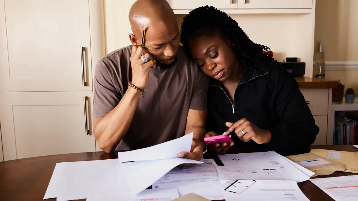 A couple sits at a table managing domestic finances, evaluating documents and using a smartphone.