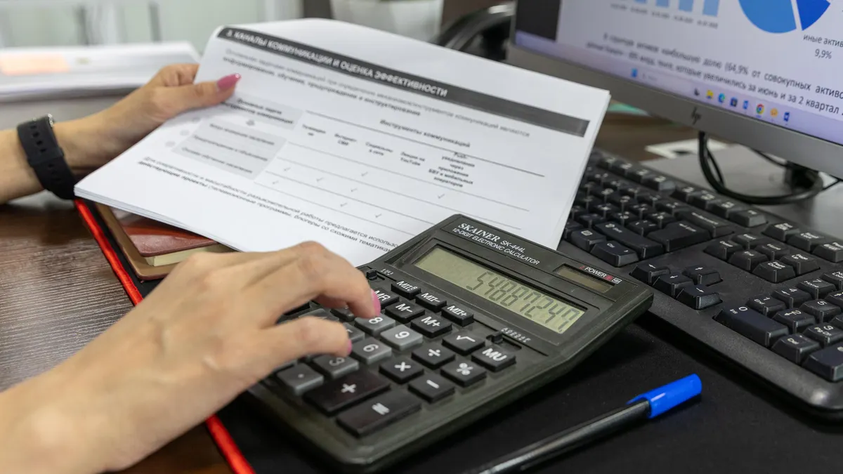 A person calculates financial data using a calculator and document, working at an office desk.