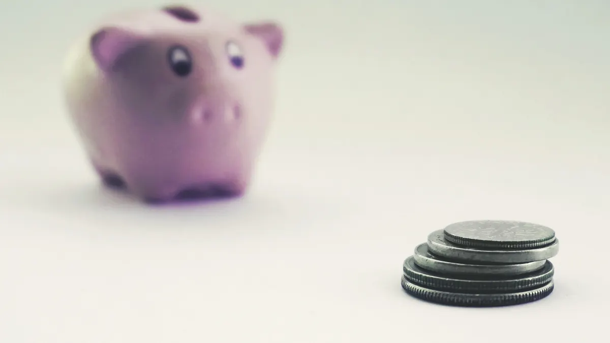 A pink piggy bank blurred in the background with stacked coins in the foreground on a white surface.