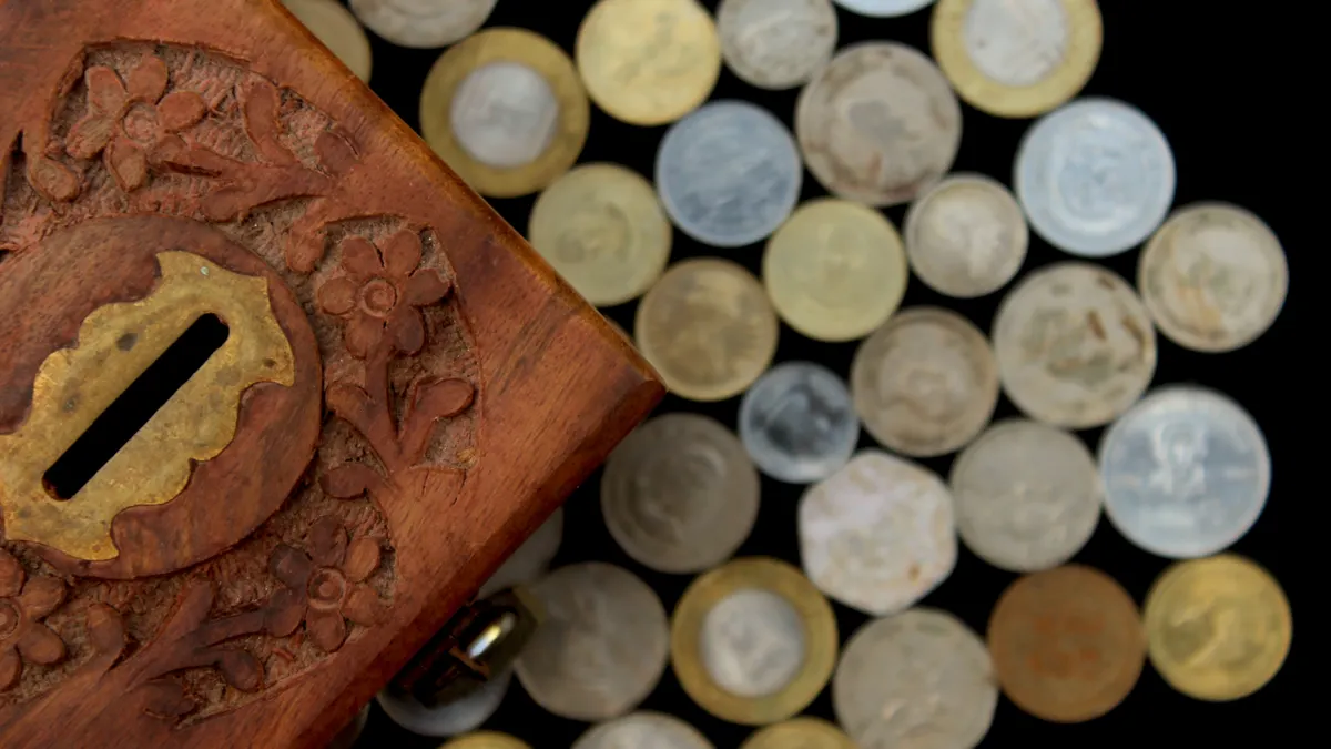 Top view of a carved wooden money box surrounded by assorted coins on a dark background.