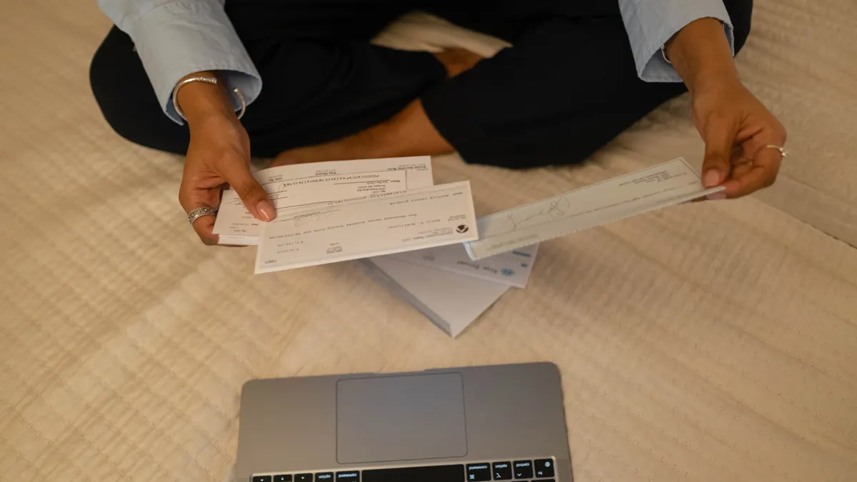 Close-up of hands holding cheques beside a laptop indoors for financial tasks.