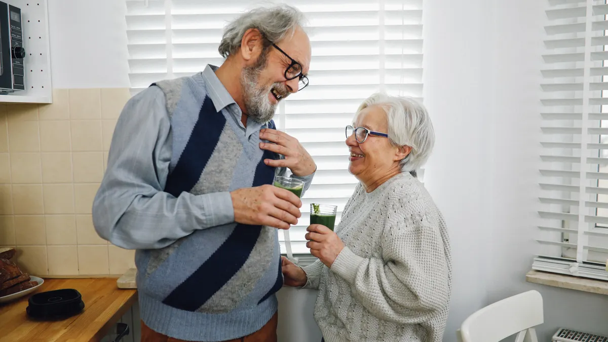 Elderly couple smiling and holding drinks in a cozy kitchen setting.