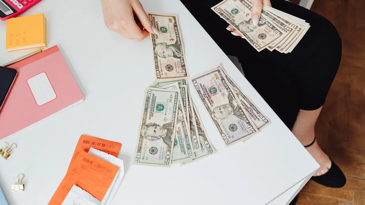 A person counting US dollar bills at a white desk with office supplies nearby.
