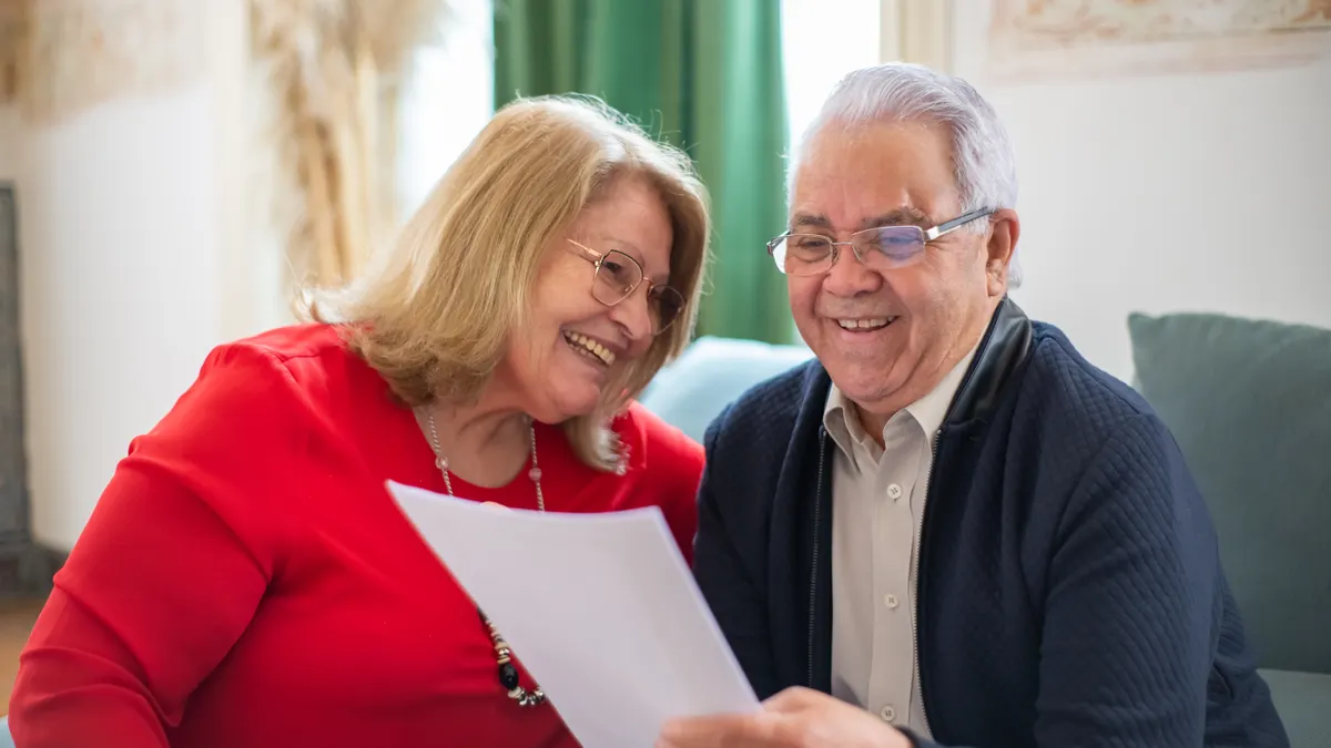 Elderly couple joyfully reading a paper together on the couch in a cozy indoor setting.