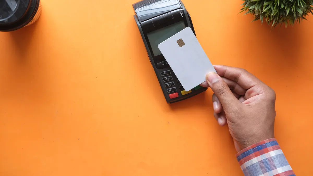 A person making a contactless payment with a credit card and card reader on a bright orange surface.